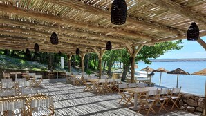 Een terras buiten met tafels en stoelen, een rieten dak en uitzicht op zee.