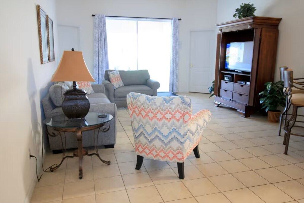A living room with a sofa, armchair, TV stand, lamp, and a glass table.