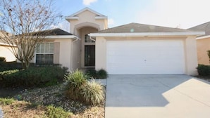 A single-story house with a garage, a front entrance with a small porch, and a landscaped front yard with shrubs and a tree. - Cozy 4 Bedroom Villa with 3 Bathrooms and South Facing Pool (CLERMONT)
