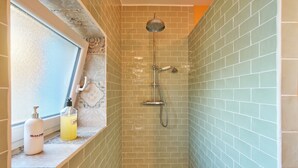 A shower with a handheld showerhead and a wall-mounted faucet, a window with a view, and a shelf with toiletries.