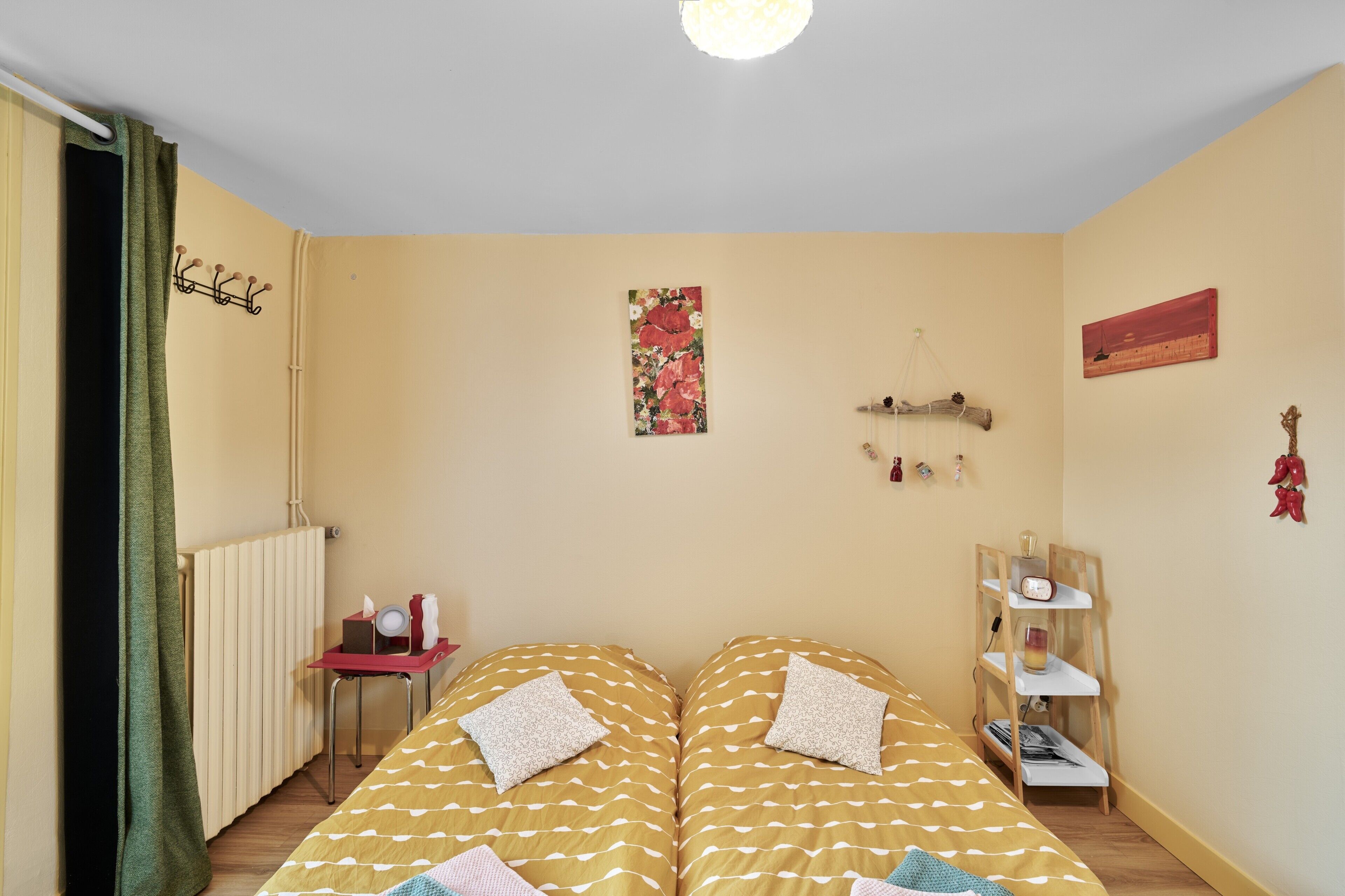 Een kamer met twee bedden, een radiator, een klein tafeltje met een rode dienblad, een plank met een klok en een schilderij aan de muur.