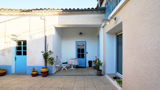 A courtyard with a tiled floor, a white wall, a blue door, a small table with a chair, and potted plants.