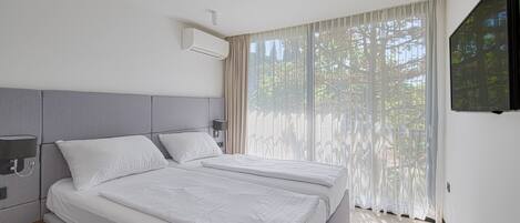 A modern hotel room with two beds, a flat-screen TV, and a large window with sheer curtains.