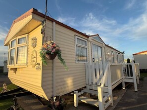 Une maison mobile dotée d’une véranda, d’un pot de fleurs suspendu et d’une boîte aux lettres bien visible.