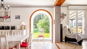 A dining area with a table and chairs, a white chair with a red cushion, a sofa with a striped cover, and a large arched window with a view of greenery.