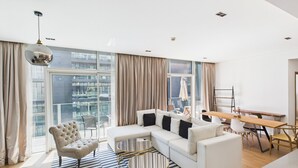 A modern living room with a large window, a sofa set, a dining table, and a chandelier. - City Walk 1BR Near Burj Khalifa Dubai Mall (Dubai)