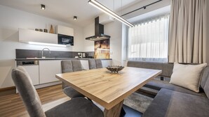 Eine moderne Küche mit eingebauten Geräten, ein Essbereich mit einem Holztisch und grau gepolsterten Stühlen sowie ein großes Fenster mit Vorhängen.