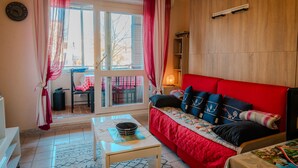 A living room with a red sofa, a small table, a window with curtains, and a view of the outside.