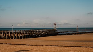 A coastal scene with a wooden pier extending into the sea, a lighthouse, and a clear sky.
