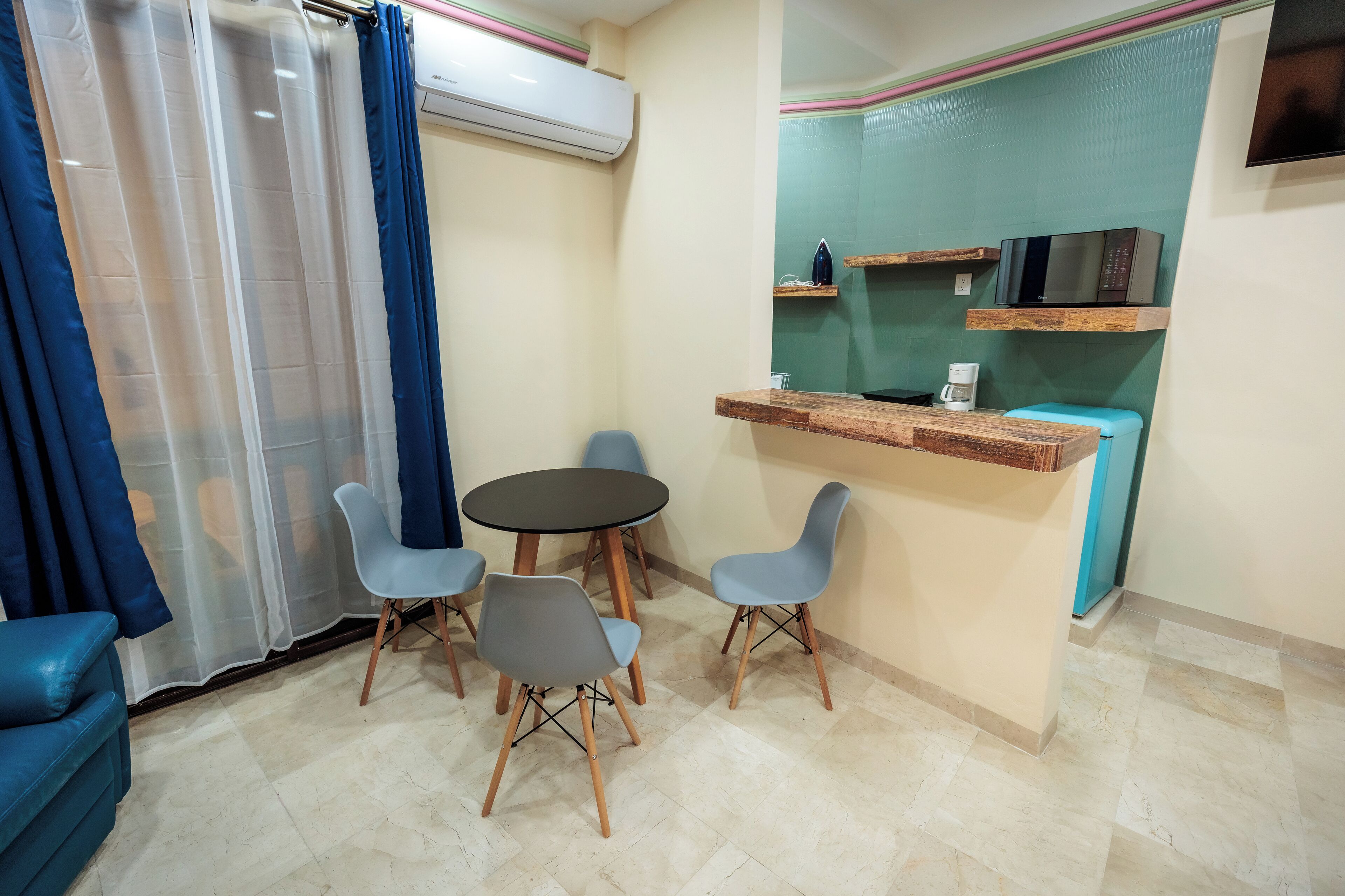 A room with a round table, four chairs, a blue sofa, a kitchen area with a microwave, and a coffee machine.