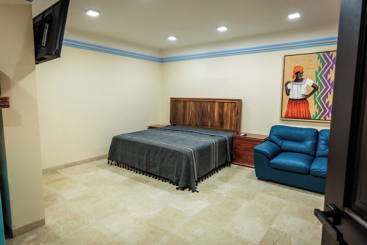 A bedroom with a bed, a blue sofa, a wooden headboard, a painting, and a television mounted on the wall.