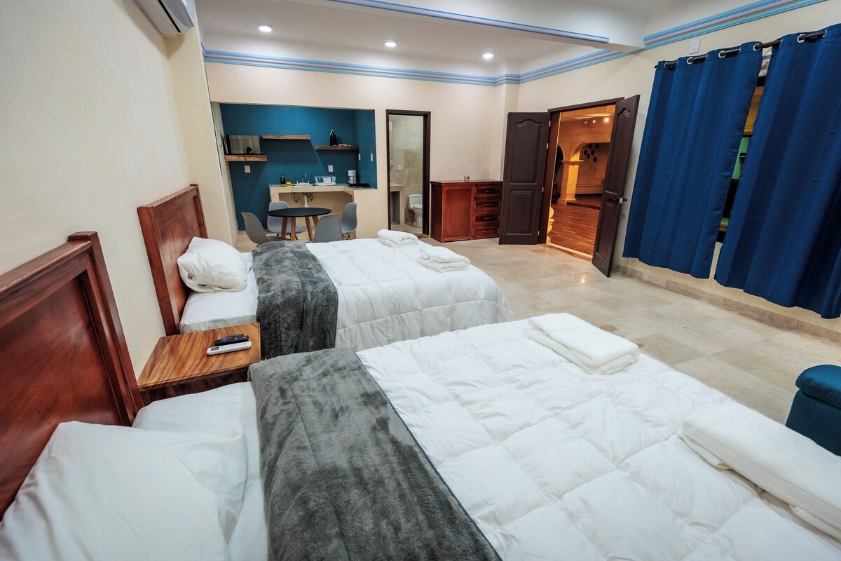 Une chambre d’hôtel avec deux lits, un bureau, une chaise, une petite table et une salle de bain visible à travers une porte ouverte.