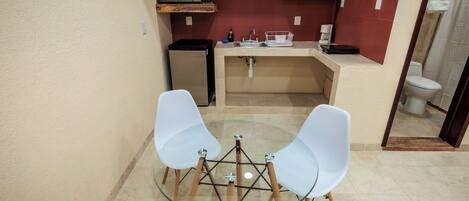A compact kitchen with a microwave, sink, and a small refrigerator. There is a glass table with two chairs in the foreground.
