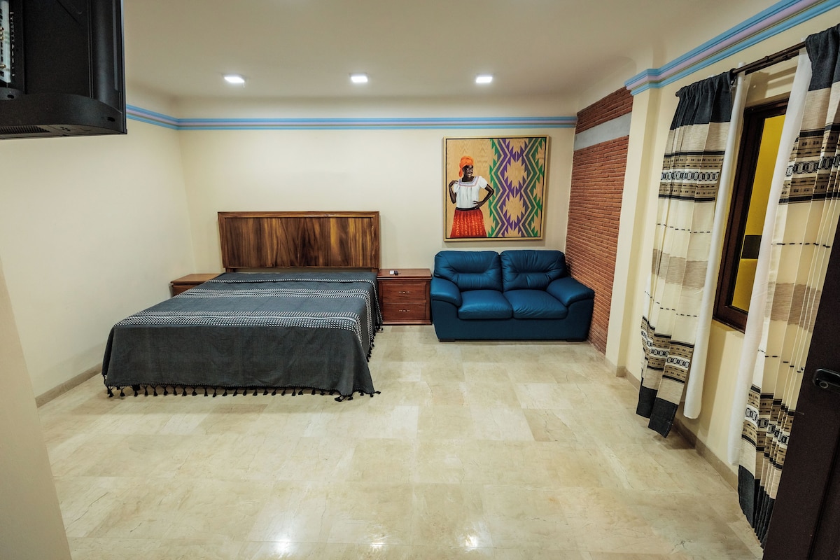 A bedroom with a bed, a blue sofa, a wooden headboard, a painting on the wall, and a patterned curtain.