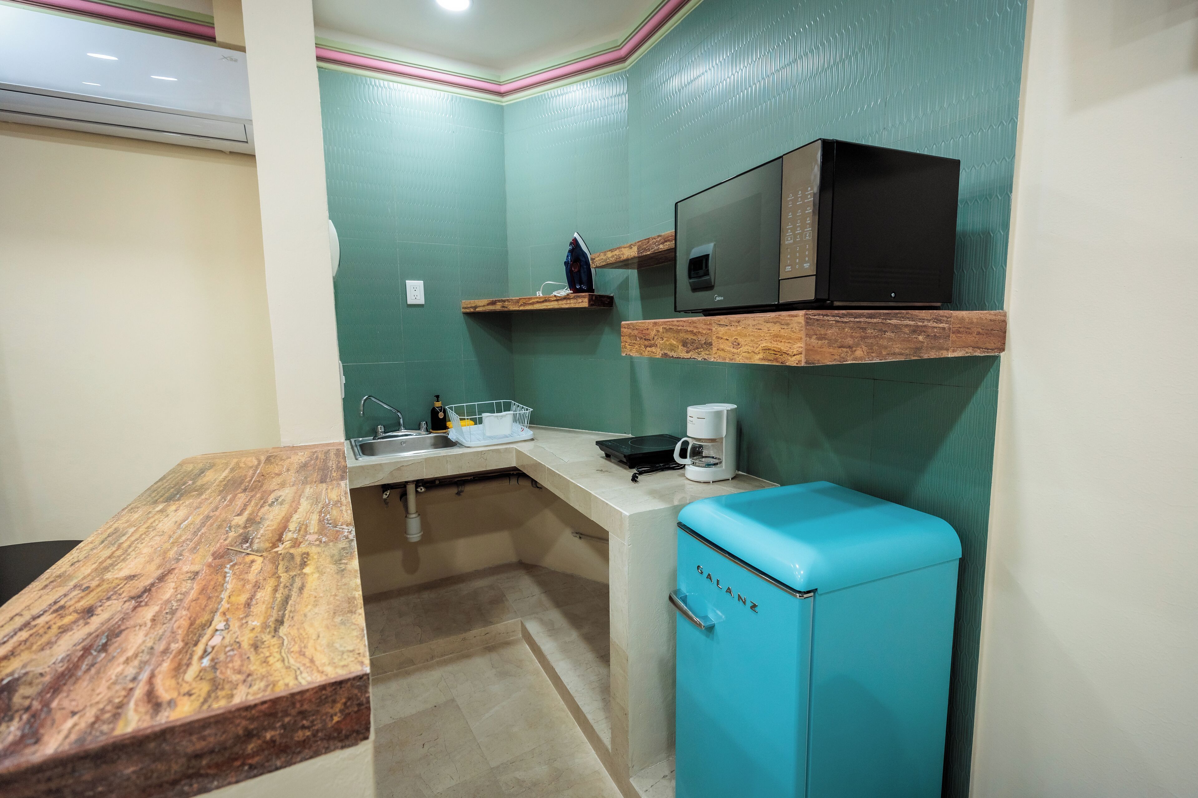 A compact kitchen with a marble countertop, a blue refrigerator, and a microwave on a wooden shelf.