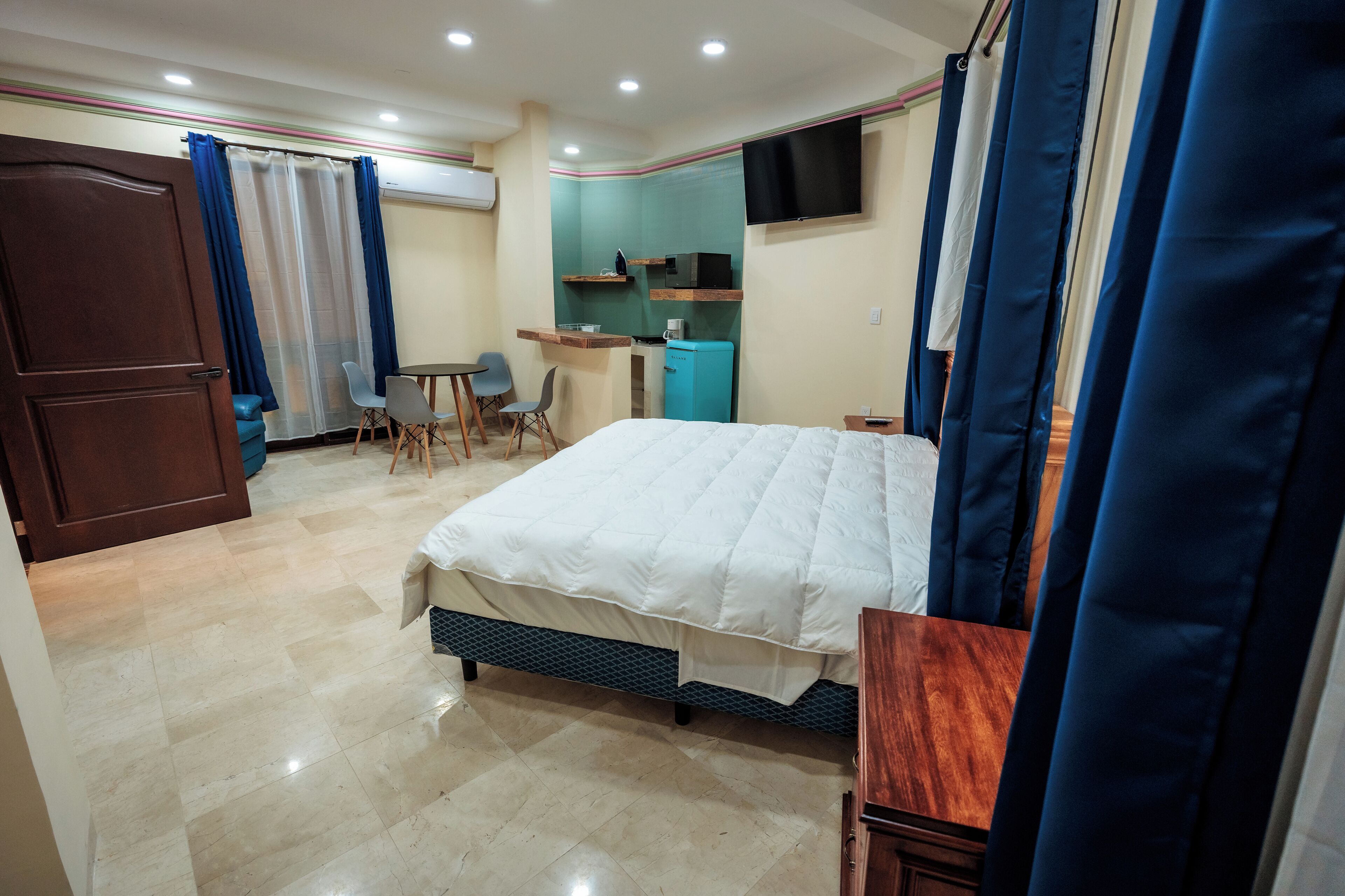 A hotel room with a bed, a desk with a chair, a small kitchenette, and a TV on the wall.
