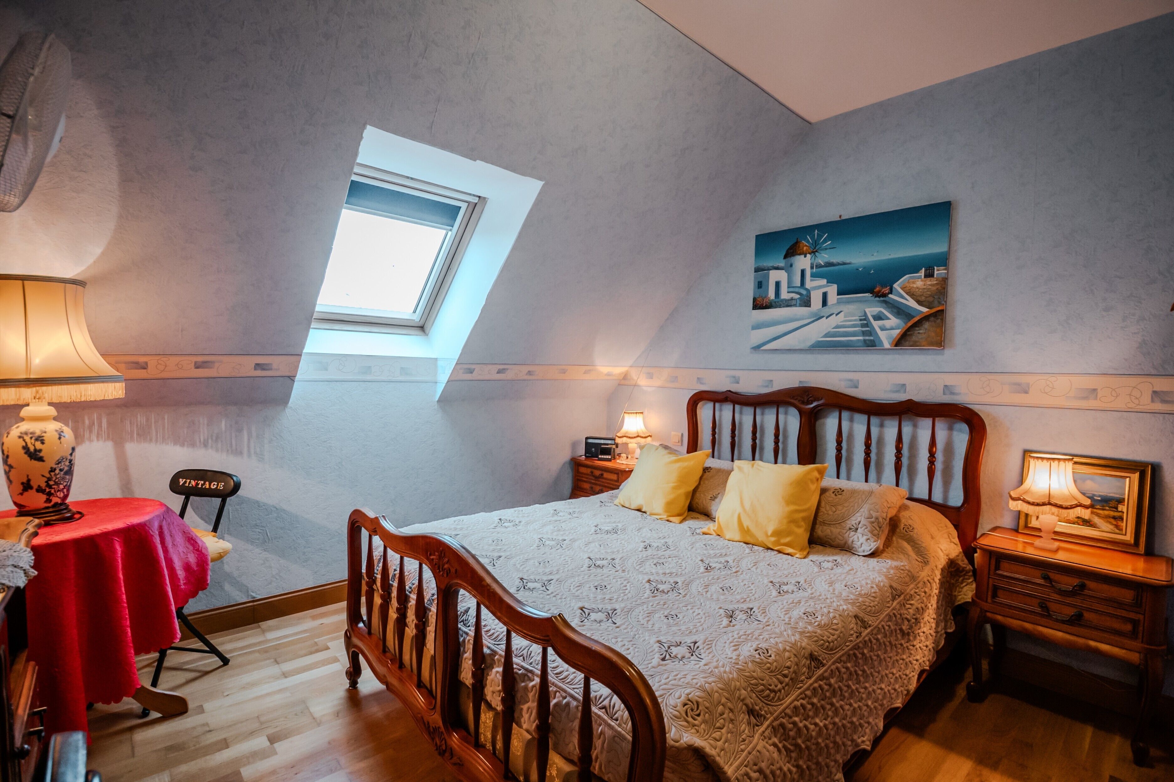 A bedroom with a skylight, a bed with a headboard, a bedside table, a nightstand, a lamp, a chair, and a table with a vase.