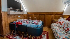 A dining area with a table set for four, a TV mounted above, and a fan on the wall. - 'Courseulles Comme À La Maison' Apartment with Private Terrace and Wi-Fi (Courseulles-sur-Mer)