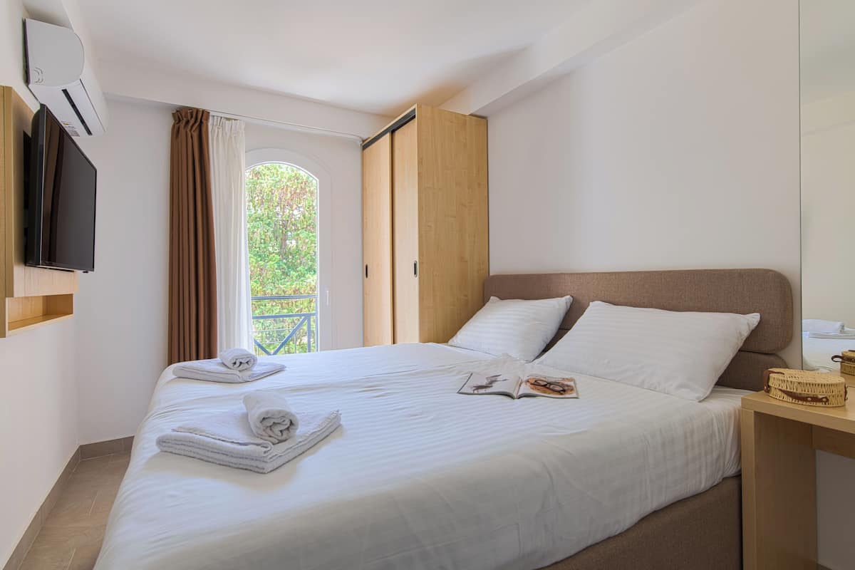 Una cama bien tendida con sábanas blancas, un televisor de pantalla plana montado en la pared y una mesita de noche de madera con una cesta.