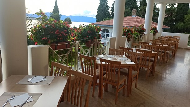 A restaurant with wooden tables and chairs, a view of mountains, and potted plants. - Hotel Delfin (Omišalj)