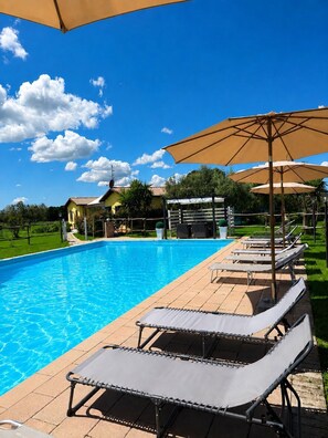 Une piscine entourée d’une pelouse et d’un bâtiment, avec des chaises longues et des parasols à proximité.