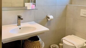 Et moderne badeværelse med hvid vask, spejl og toilet. To kurve står på gulvet, og en skraldespand er placeret ved siden af toilettet.