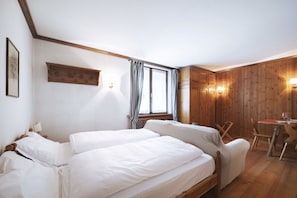 A bedroom with two beds, a wooden wardrobe, a desk with a chair, and a window with curtains.