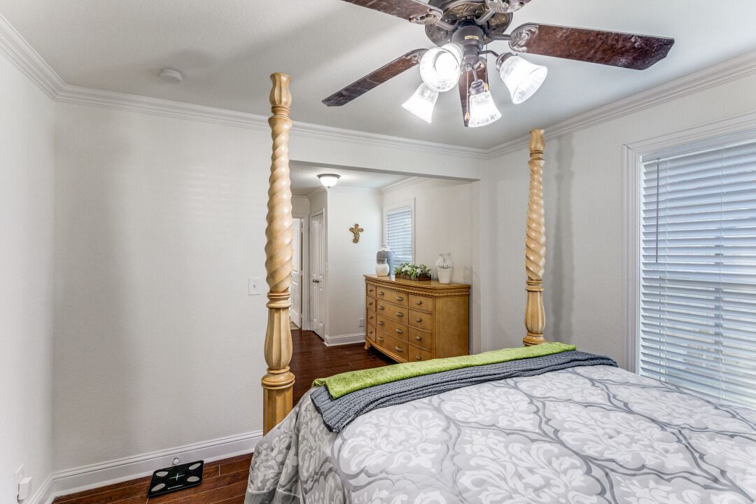 A bedroom with a four-poster bed, a ceiling fan, a dresser, and a window with blinds.