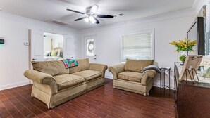 A living room with two beige couches, a ceiling fan, hardwood flooring, and a wooden cabinet with a vase of flowers. - Charming house in Fort Worth walking distance to TCU (Fort Worth)
