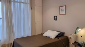 Une chambre à coucher avec un lit, une table de chevet, une lampe, un radiateur et une fenêtre avec des rideaux.