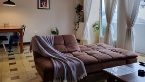 Un salon avec un canapé marron, une table basse en bois, une table à manger avec des chaises, un luminaire suspendu, un tableau encadré au mur et une fenêtre avec des rideaux.