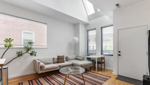 Una sala de estar moderna con un sofá, una mesa de centro de vidrio y un tapete a rayas. Hay dos ventanas con persianas, una puerta y una pequeña mesa con una silla.