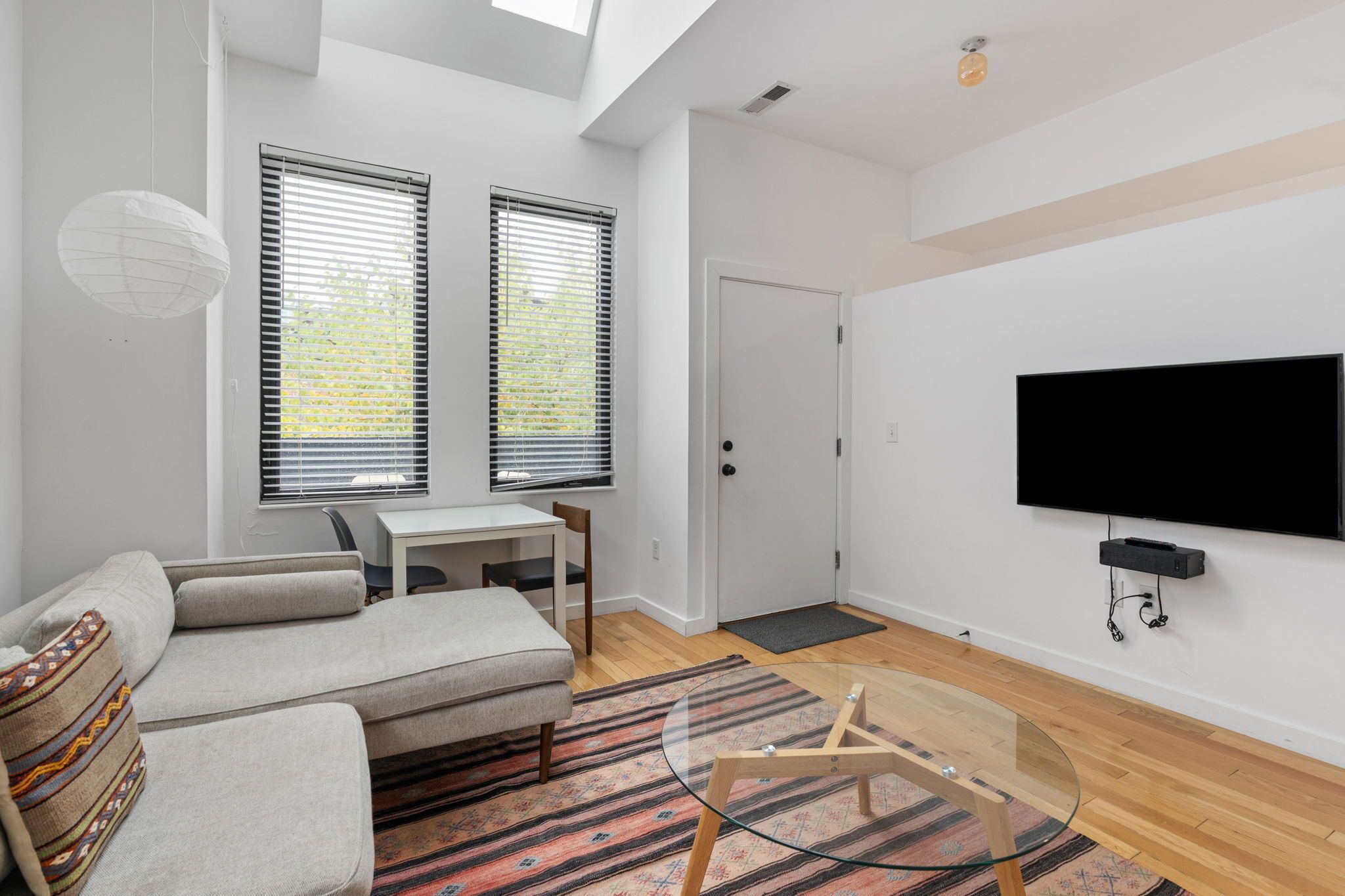 Un salon moderne avec un canapé, une table basse en verre, un téléviseur fixé au mur et deux fenêtres équipées de stores.