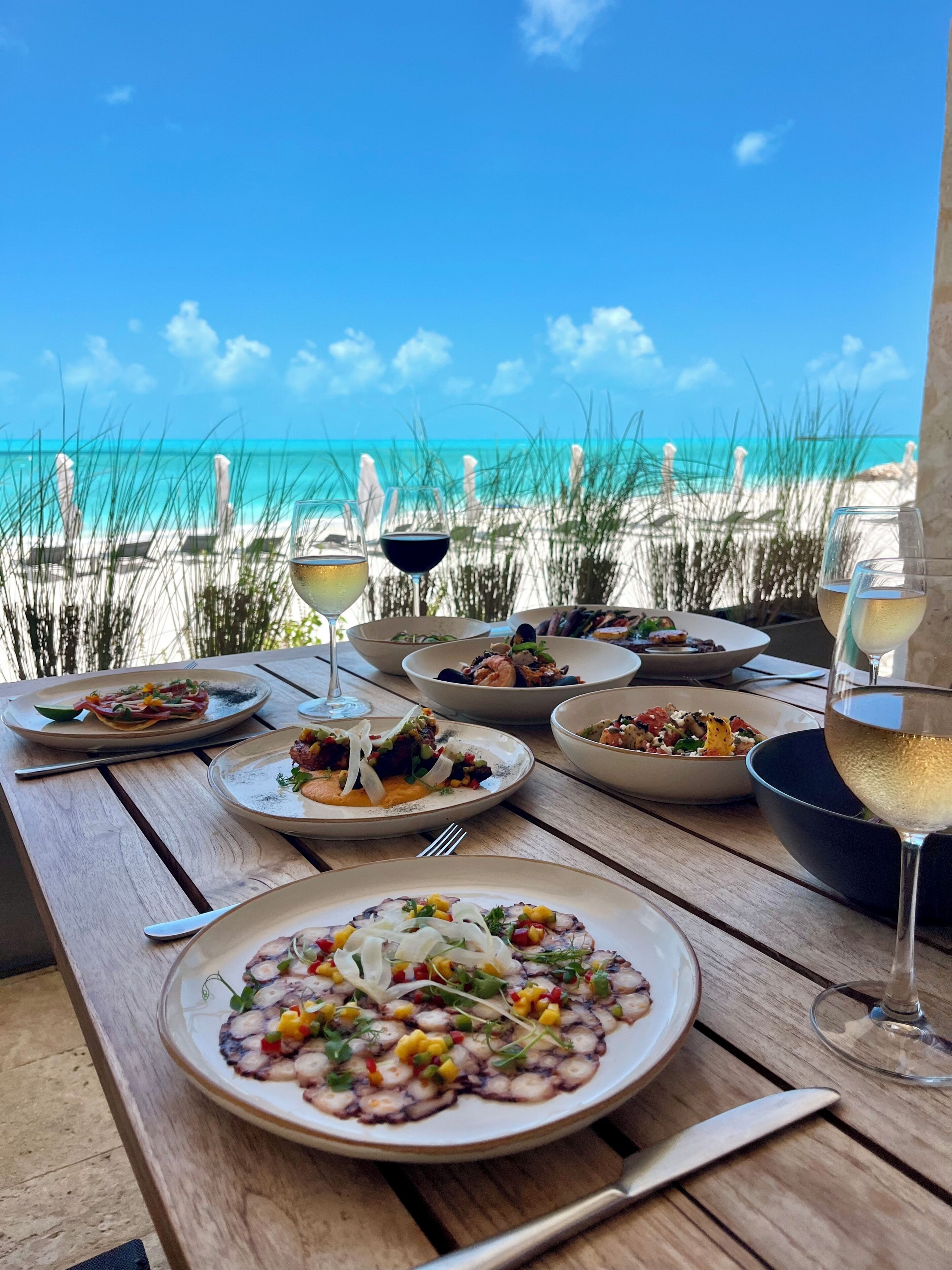 Une table dressée avec différents plats et des verres de vin, donnant sur une plage aux eaux d’un bleu limpide et au sable blanc.