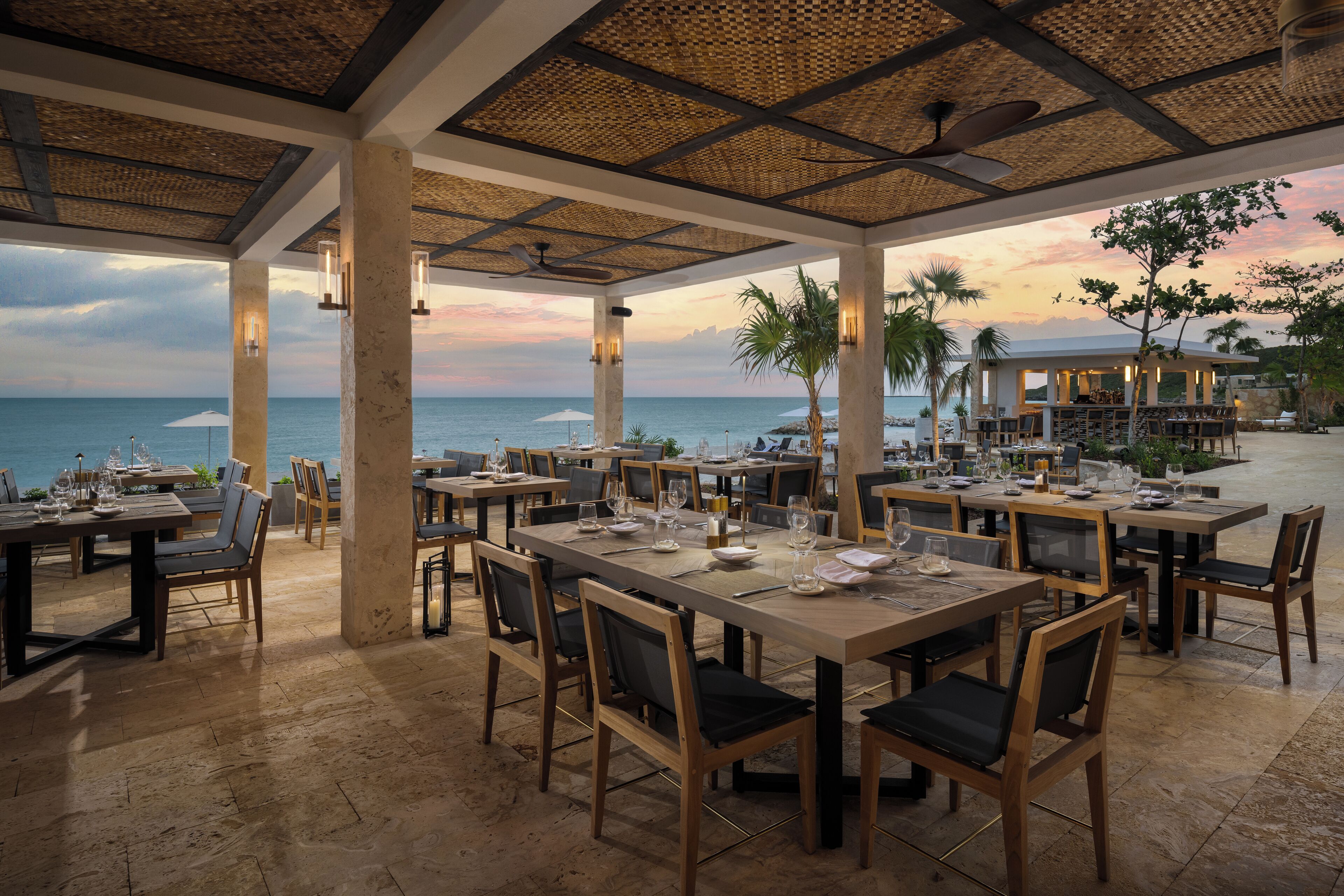 A restaurant with outdoor seating overlooking the sea, featuring wooden tables and chairs, and a thatched roof structure.