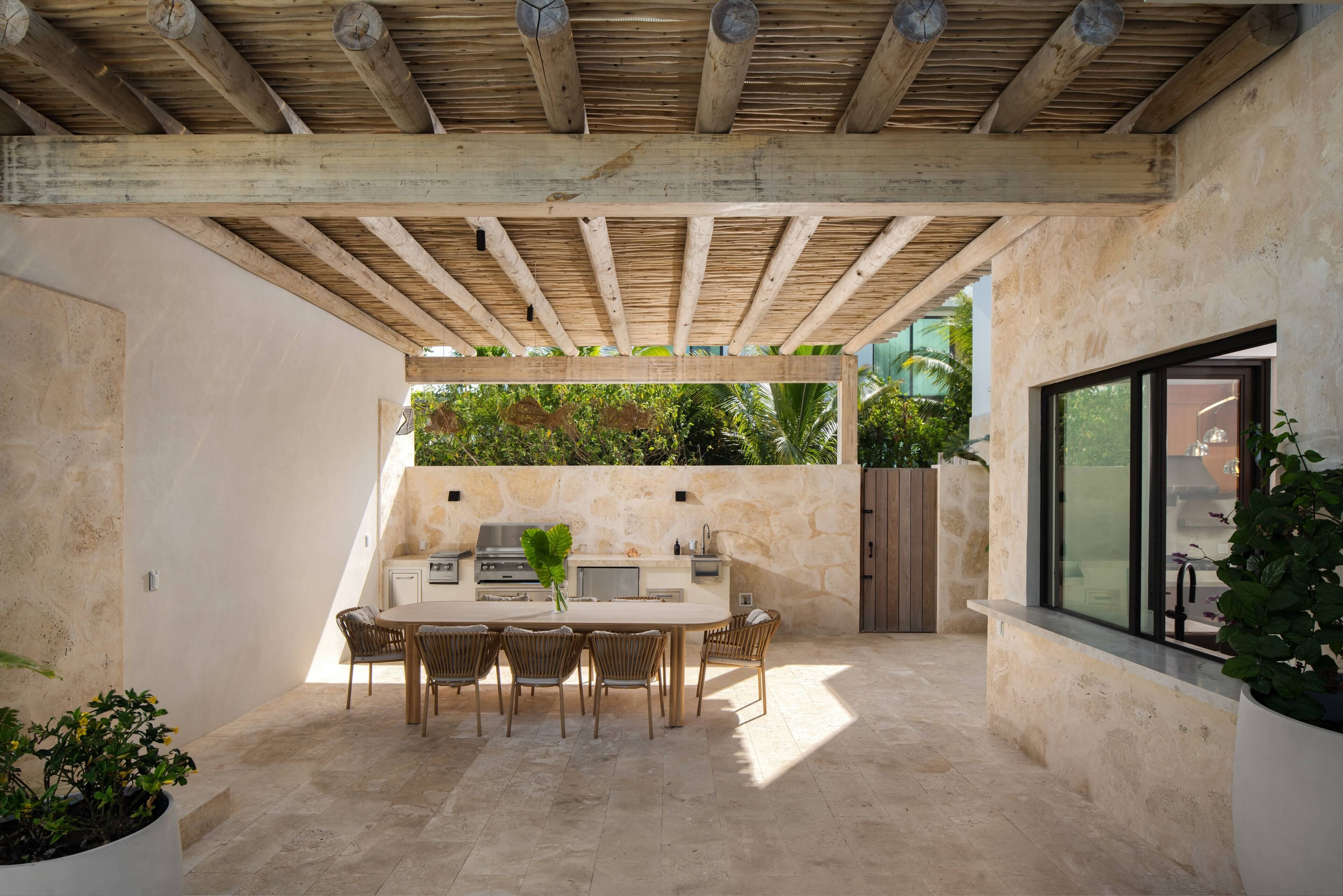 Une terrasse extérieure dotée d’une pergola en bois, d’une table à manger avec des chaises, d’un barbecue et d’un mur en pierre agrémenté de verdure.