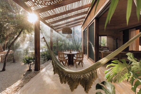 A hammock under a wooden pergola, a round table with chairs, and a view of tropical vegetation.