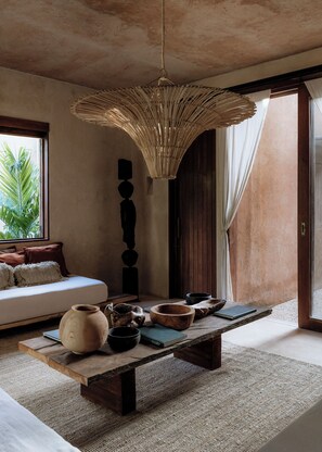 A room with a wooden bench, a hanging light fixture, a sofa, and a view of greenery outside.