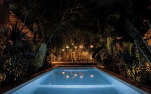 A nighttime pool area with a clear pool, surrounded by tropical plants and illuminated by hanging lanterns.