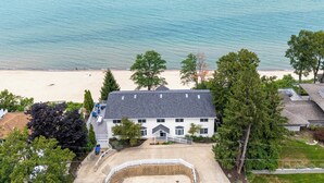 A white two-story house with a grey roof, a front porch, and a driveway leading to a white picket fence. - New Buffalo Sandcastle (New Buffalo)