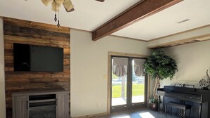 A living room with a fireplace, a television mounted on a wooden wall, a dining area with a piano, and a large window with curtains.