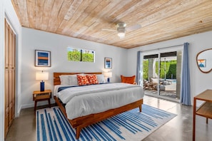 A bedroom with a wooden ceiling, a large bed, two bedside tables with lamps, a view through a glass door to an outdoor area, and framed artwork on the walls.