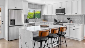 A modern kitchen with white cabinetry, a stainless steel refrigerator, a built-in oven, and a large island with bar stools. - Sunny Dreams - Mid-Century Pool & Spa Retreat (Palm Springs)