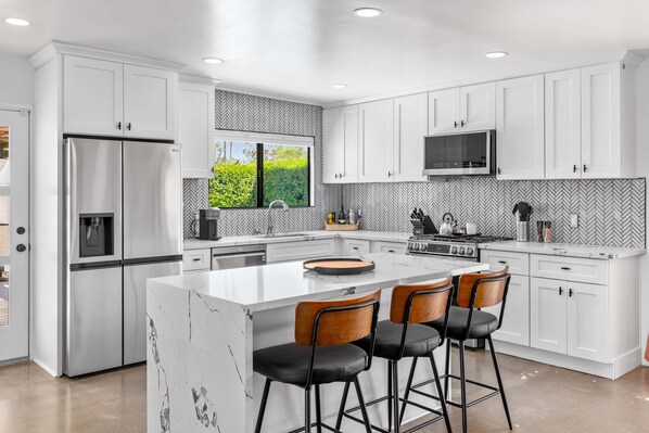 A modern kitchen with white cabinetry, a stainless steel refrigerator, a built-in oven, and a large island with bar stools.