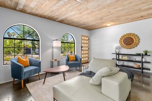 Een moderne woonkamer met een houten plafond, grote rondboogvensters, een witte bank, twee fauteuils, een klein tafeltje en een zwarte boekenkast.
