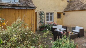 A courtyard with a yellow building, a wooden gate, a small garden with flowers, and outdoor furniture. - The Old Dairy (Chippenham)