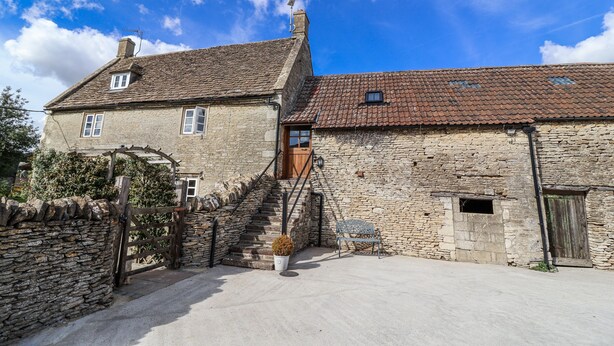 A stone building with a wooden staircase leading to a door, a small window, and a stone wall with a gate. - The Old Dairy (Chippenham)