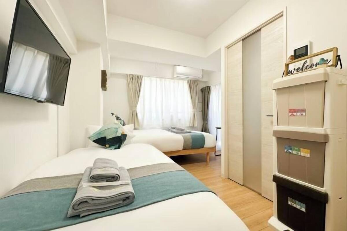 Een hotelkamer met twee bedden, een aan de muur bevestigde televisie, een airconditioningapparaat en een 'Welcome' bord op de deur.