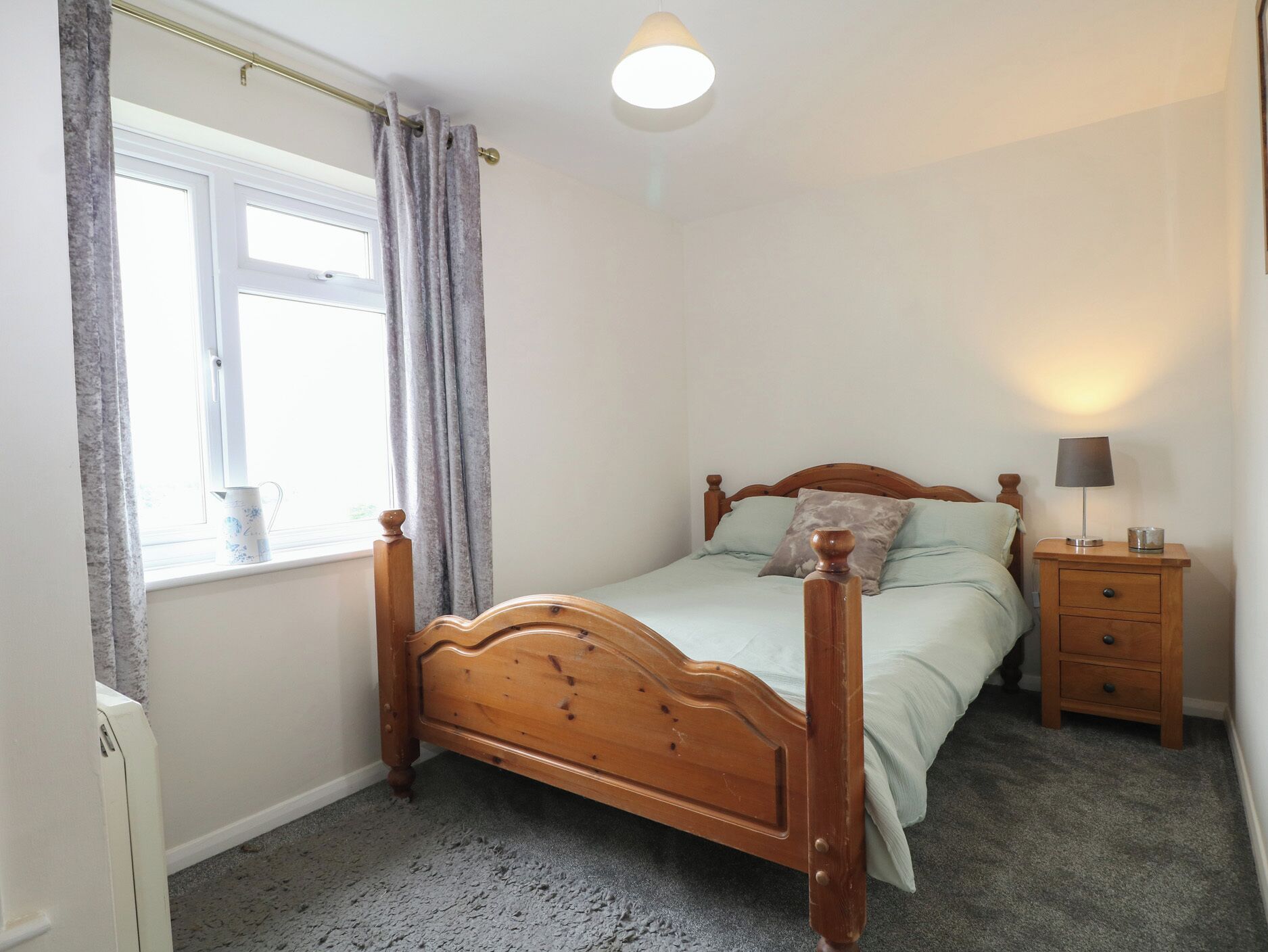 A bedroom with a wooden bed, a bedside table with a lamp, a window with curtains, and a carpeted floor.