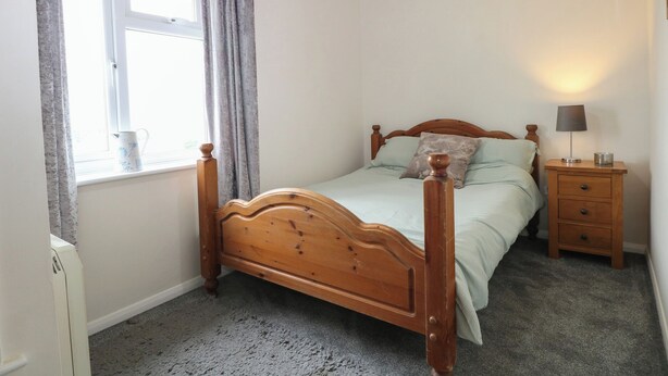 A bedroom with a wooden bed, a bedside table with a lamp, a window with curtains, and a carpeted floor.